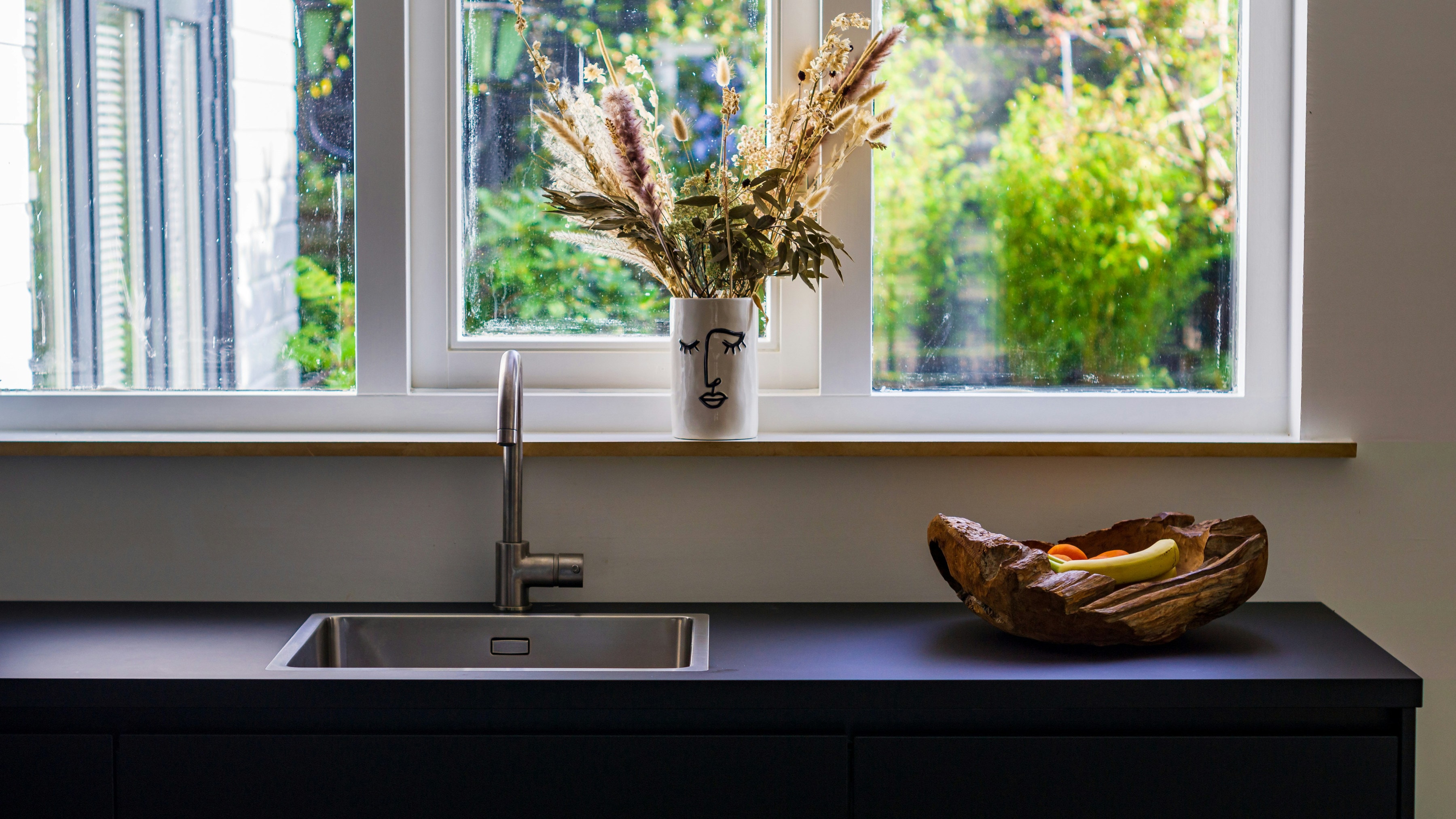 Stainless steel sink and tap on a matt black worktop below a garden window with dried flowers