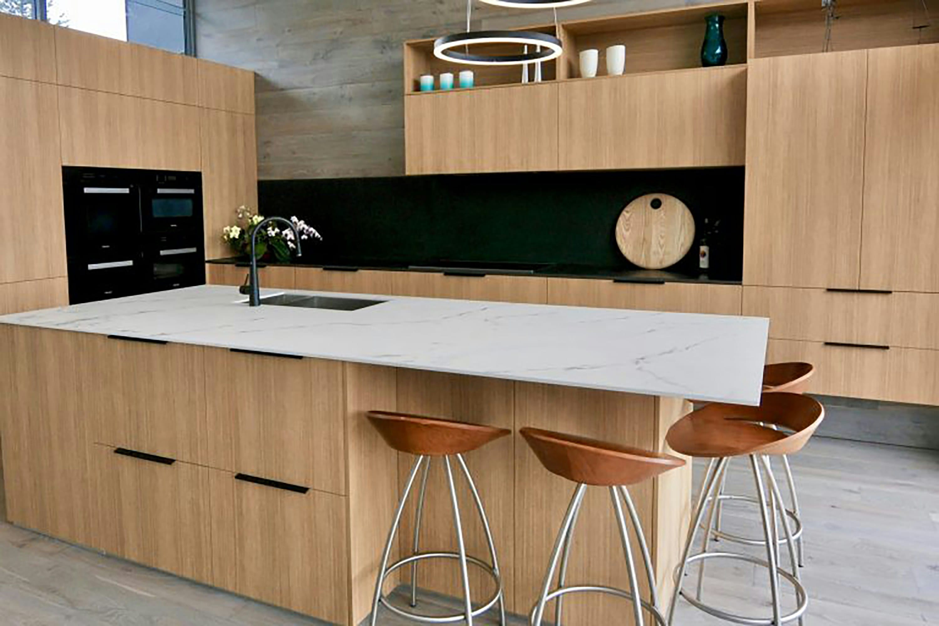 Light oak island with thin marble look top, black splashback, black wall ovens and copper stools